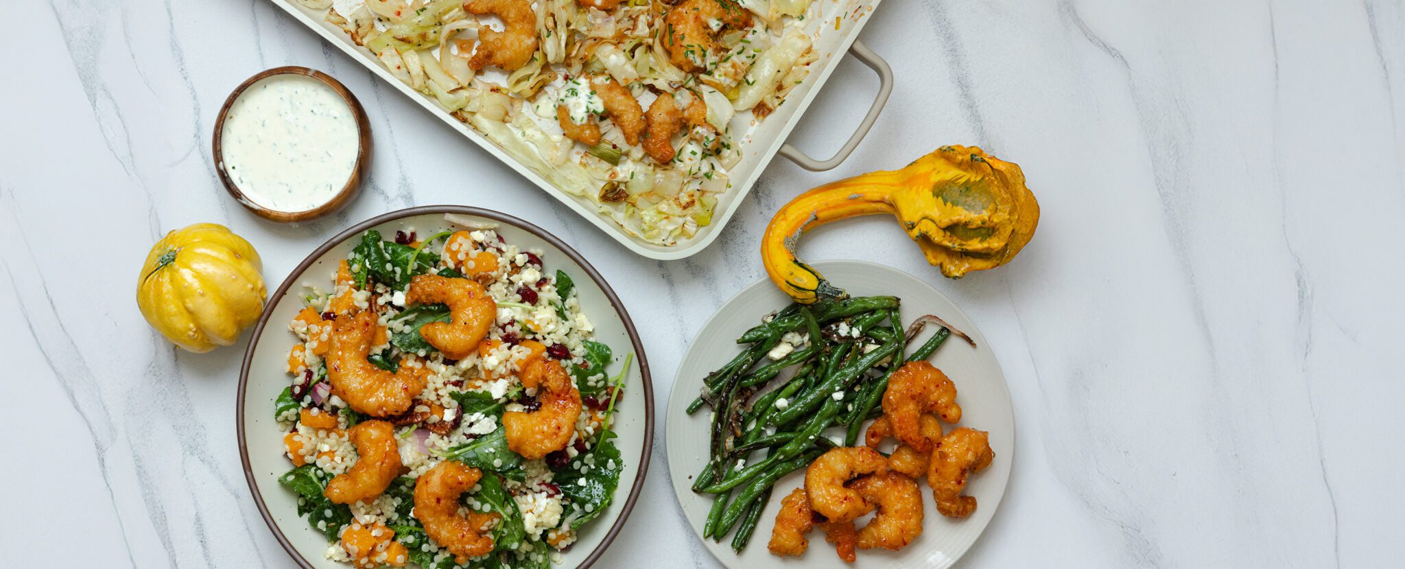 Overhead view of several dishes including a Fall Couscous Salad with shrimp, a casserole dish of Sweet & Spicy Shrimp with Cabbage, and a plate of shrimp with roasted green beans. All served on a marble tabletop with a side of chive aioli and two autumnal pumpkins.