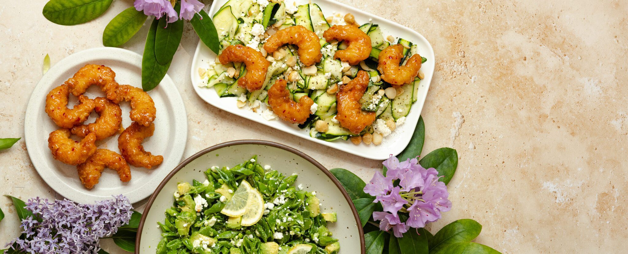 Overhead view of tablescape with Social Kitchens Sweet & Spicy Shrimp, Shaved Zucchini Salad, and Snap Pea & Avocado Salad. Arranged with purple flowers and green foliage surrounding the three plates.