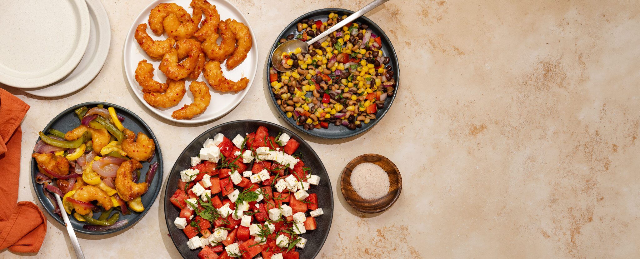 Overhead view of an assortment of dishes including Social Kitchens Sweet & Spicy Shrimp, Cowboy Caviar, and Watermelon Feta Mint Salad. Off to the side are additional white plates, an orange cloth napkin, and a small wooden bowl of salt.