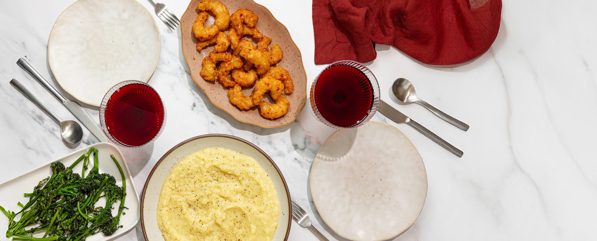 Overhead view of a tablescape containing miso butter mashed potatoes, broccolini, Social Kitchens Sweet & Spicy Shrimp, glasses of wine and a set of tableware all over a marble table surface.