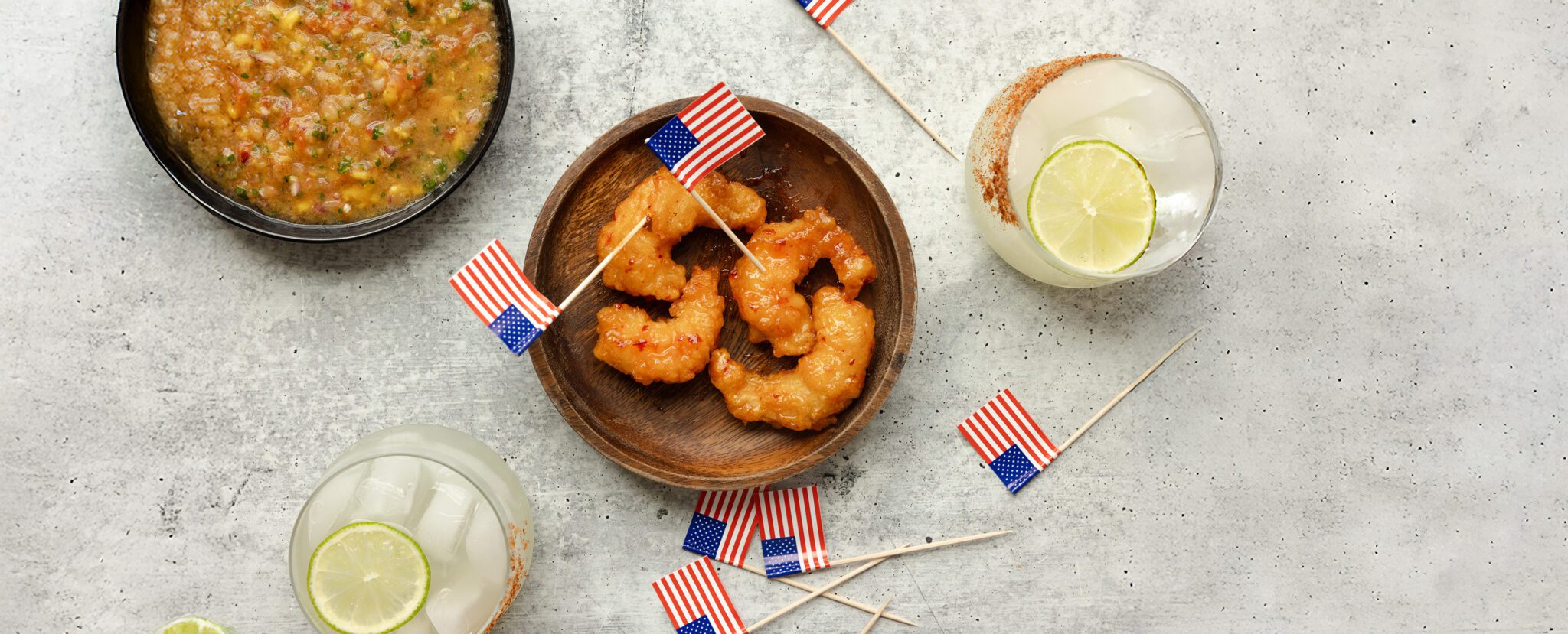 Overhead view of a wooden bowl of Social Kitchens Sweet & Spicy Shrimp, a black bowl of mango salsa, two glasses of drinks with lime slices and tajin coated rims. Scattered throughout the tablescape are small paper American flags on toothpicks.