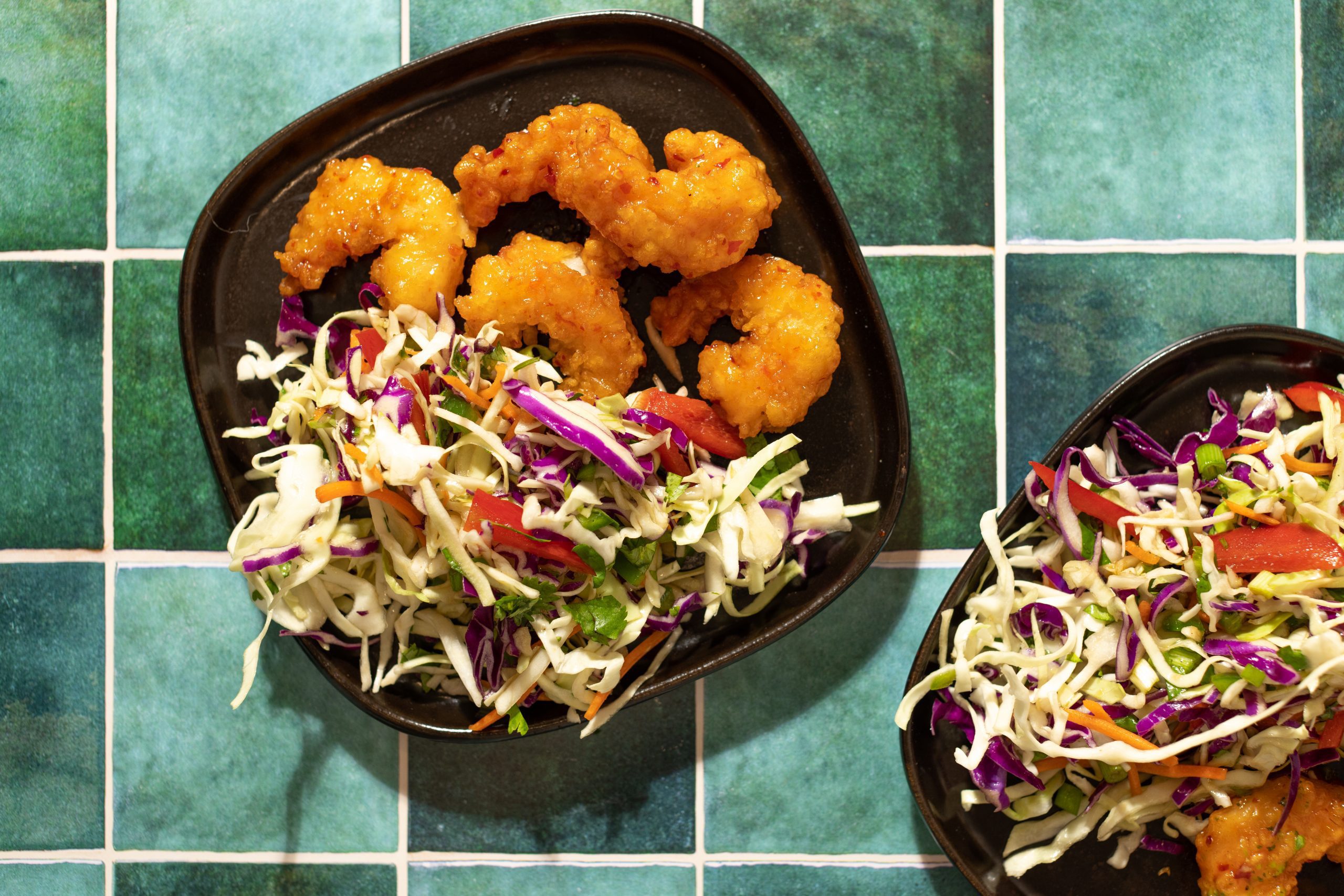 Asian inspired slaw served with Social Kitchens Sweet & Spicy Shrimp on rounded square plates over a green tile surface tabletop.