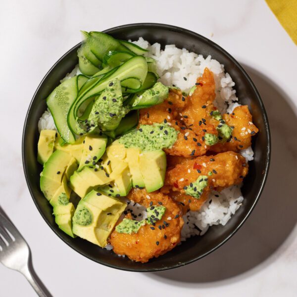 A black bowl filled with Social Kitchens Sweet & Spicy Shrimp, rice, thinly sliced cucumber ribbons, cubed avocado pieces, and black sesame seeds for garnish.