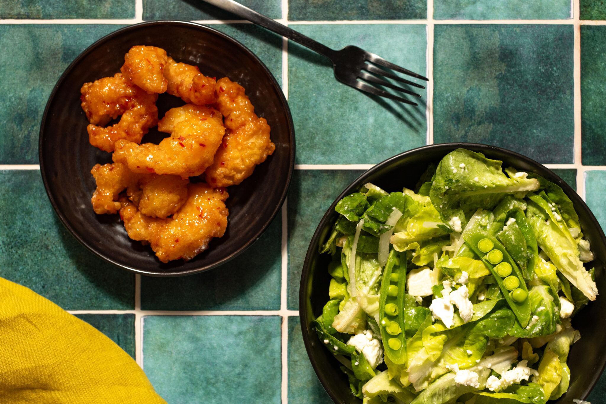 Herby spring pea salad on a black plate next to another black plate of Social Kitchens Sweet & Spicy Shrimp. Green-tiled surface with a black fork and yellow cloth napkin.