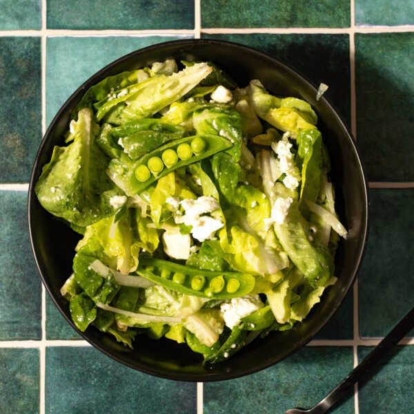 Overhead view of an herby spring pea salad served in a black bowl with a black fork over a green-tiled surface.