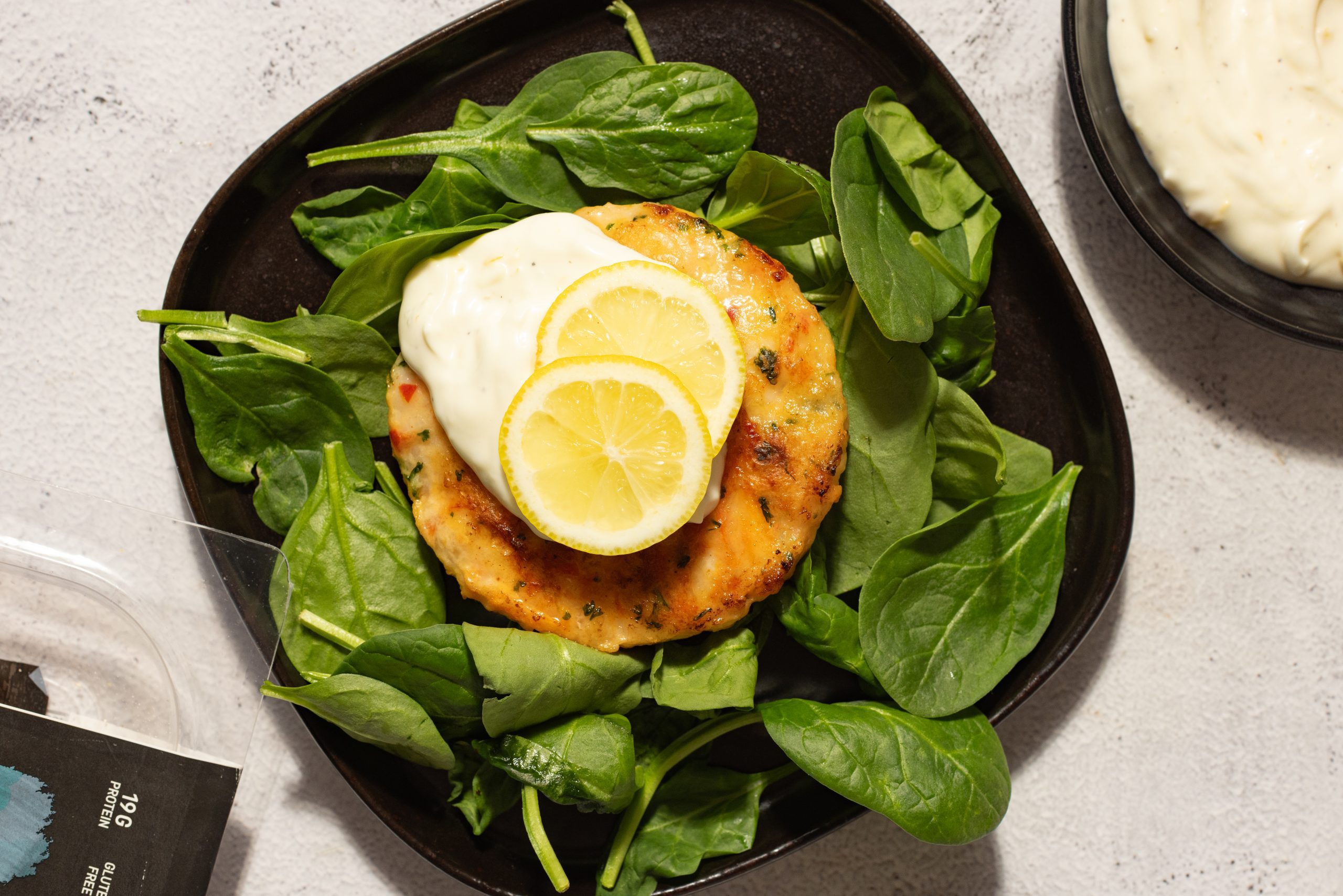 A Social Kitchens premium shrimp burger with lemon garlic aioli and 2 lemon slices, all served over a bed of spinach on a black plate. Cropped off the edge of the image is a bowl of aioli and burger packaging.