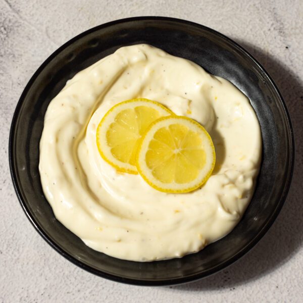 A black bowl on a neutral colored surface of lemon garlic aioli spread garnished with 2 slices of lemon.