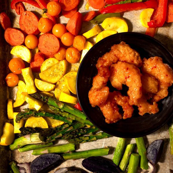 Metal sheet pan of colorful vegetables arranged by colors of the rainbow. Veggies include (from bottom to top) purple baby potatoes, sliced red onions, asparagus, yellow squash, orange cherry tomatoes, carrots, and red bell peppers. Included on the pan is a black plate of Social Kitchens Sweet & Spicy Shrimp.