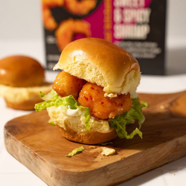 Social Kitchens Sweet & Spicy Shrimp slider burger with lettuce and mayo served on a wooden cutting board. Blurred background features box of Social Kitchens Sweet & Spicy Shrimp box packaging and empty slider buns on a white tabletop surface.