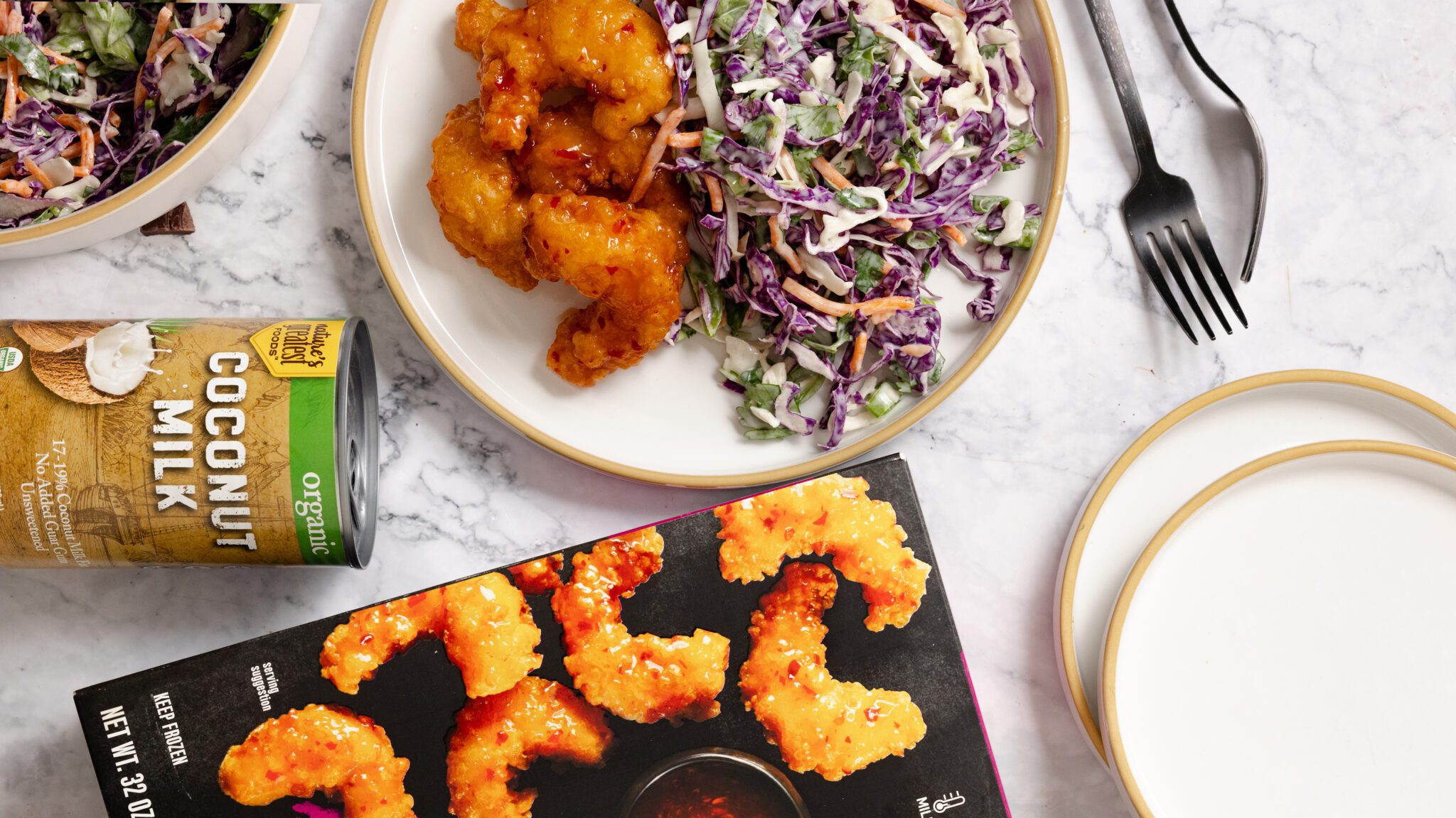 Overhead view of Social Kitchens Sweet & Spicy Shrimp box, can of coconut milk, and a plate of coconut slaw and sweet & spicy shrimp, all over a white marble surface.