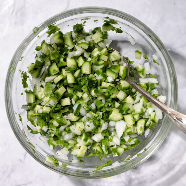 Overhead view of a clear bowl filled with diced cucumber, cilantro, white onion, and jalapeño with a metal spoon resting inside. Bowl is resting over a white marble surface.