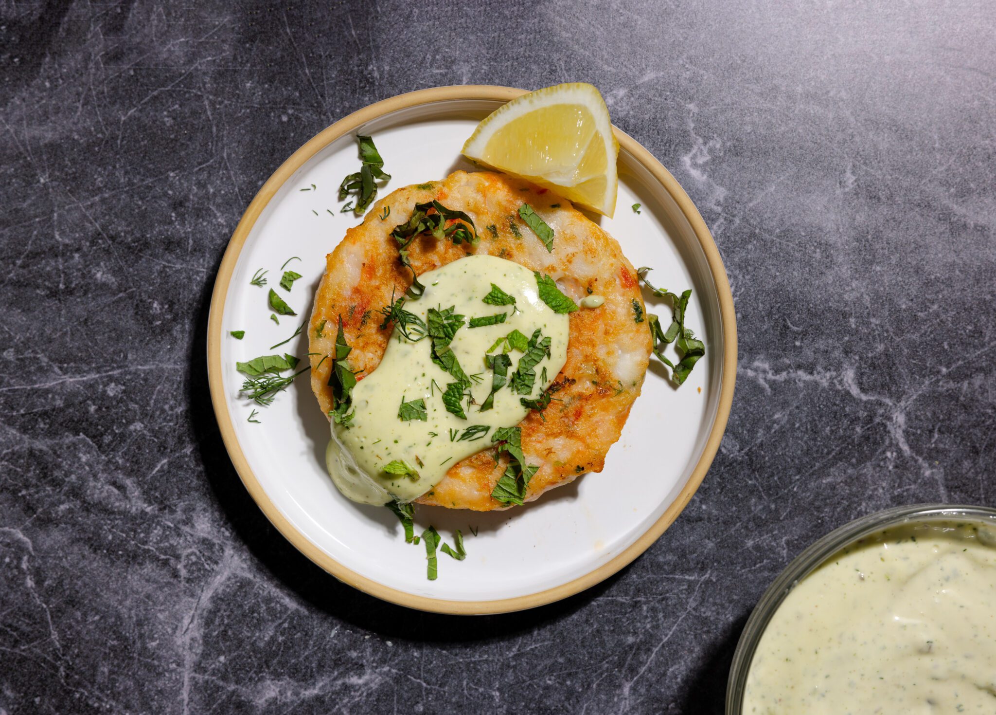 On a dark gray marble surface, an overhead view of a Social Kitchens Premium Shrimp Burger patty with herby aioli dip, herb garnishes, and a lemon wedge. In the lower left cropped off the image is a separate bowl of herby aioli.