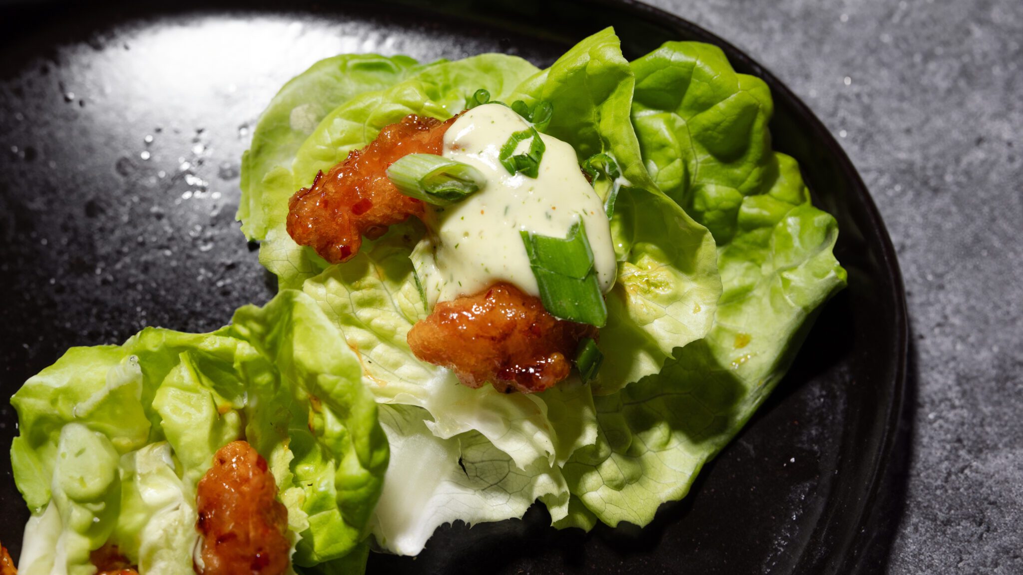 Overhead view of lettuce topped with Social Kitchens Sweet & Spicy Shrimp, herby aioli, and green onions served on a black plate.