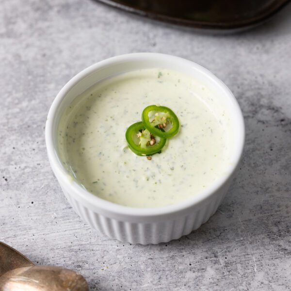 White bowl of spicy jalapeño aioli, garnished with two jalapeño slices. Served over a white gray textured surface.