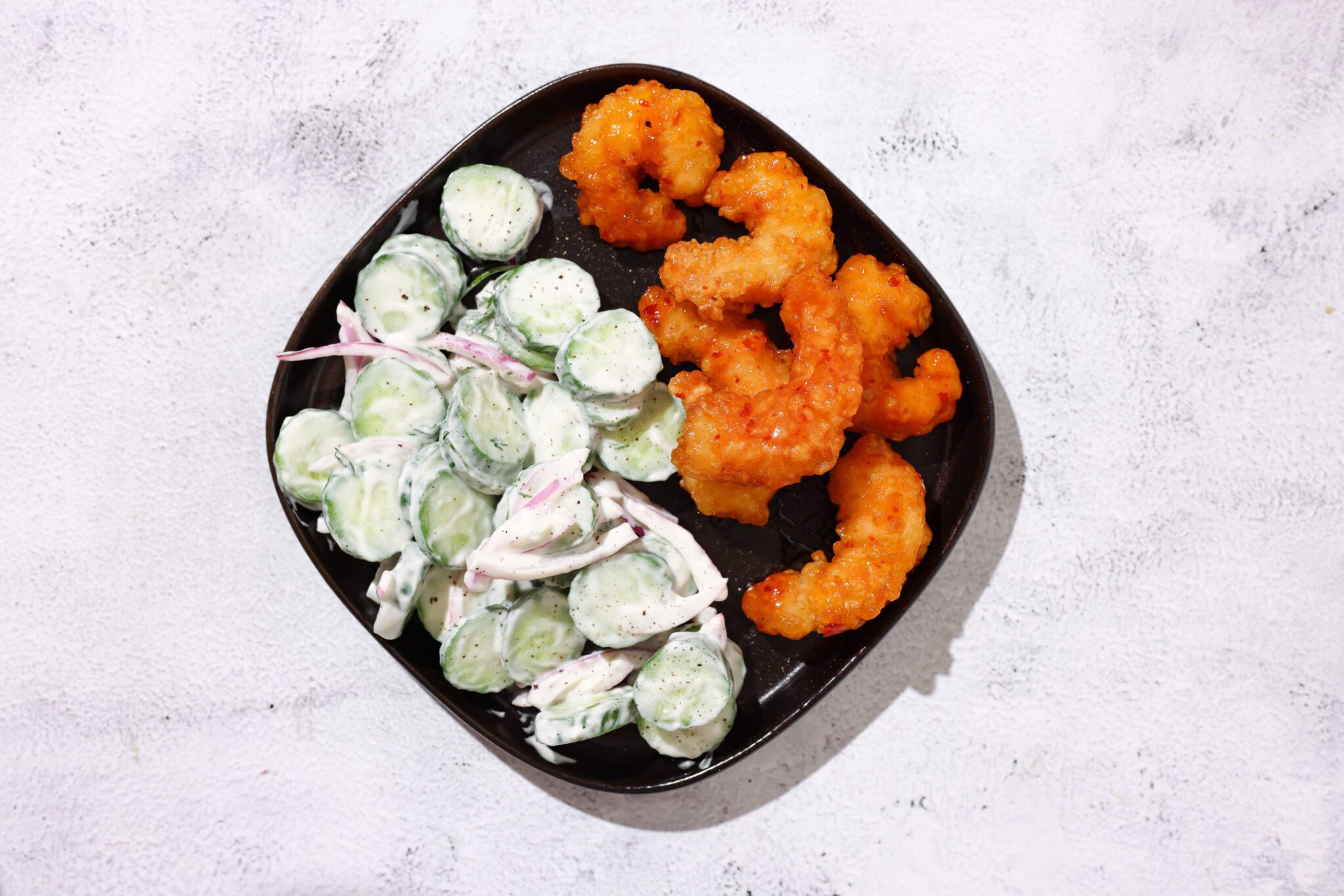 Overhead view of sweet & spicy shrimp and a yogurt cucumber salad on a black plate, served over a white marble surface.