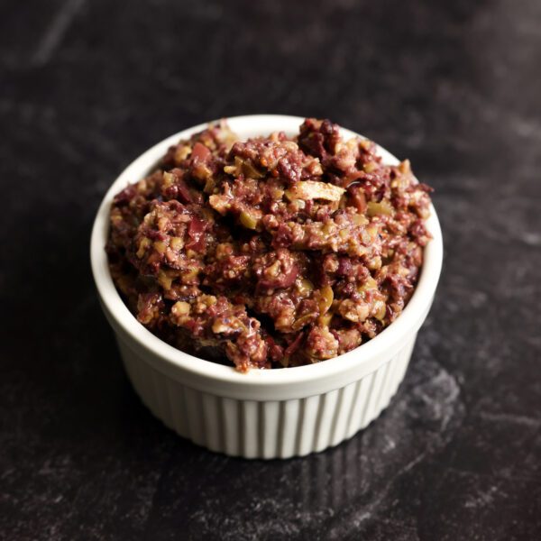 Three-quarter view of a white ramekin filled with olive tapenade, served over a black marble surface.