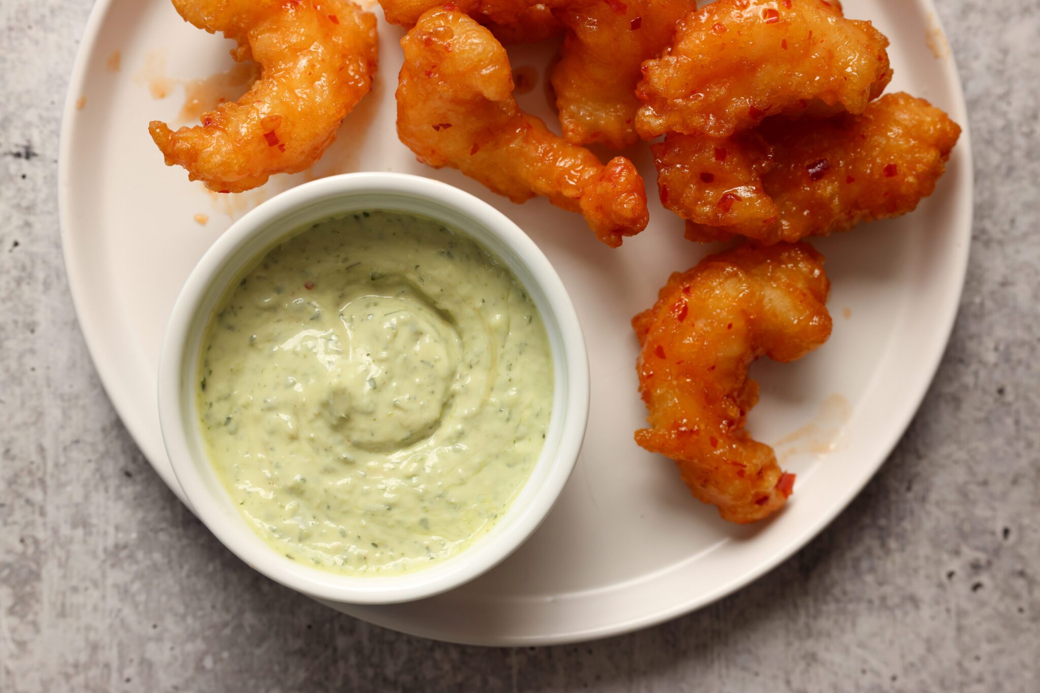 Overhead view of a white plate with sweet & spicy shrimp and a side bowl of pesto aioli, placed on a white textured surface.