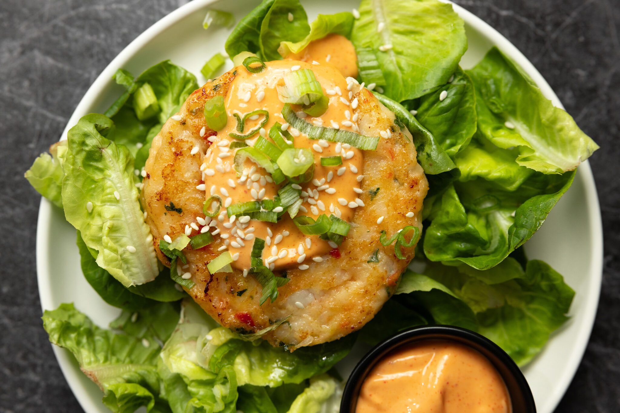 Overhead view of a Social Kitchens Premium Shrimp Burger patty over a bed of lettuce greens with a side dish of ginger gochujang aioli sauce all on a white plate served over a black marble tabletop.