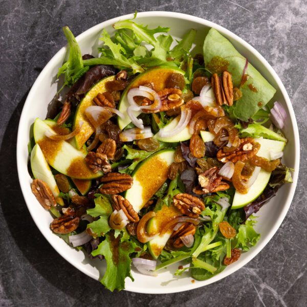 Overhead view of a round white plate of salad greens, green apples, pecans, shallot slices, and dressing, all served on a black marble surface.