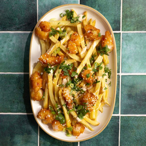 Overhead view of a large white platter filled with loaded sweet & spicy shrimp fries consisting of french fries, shrimp, cilantro, green onion, and sesame seeds. Served over a textured green title surface.