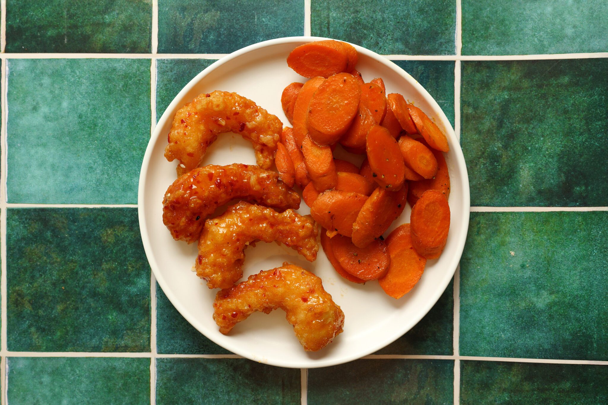 Overhead view of a white plate full of sweet & spicy shrimp and maple roasted carrot slices. Served over a textured green title surface.