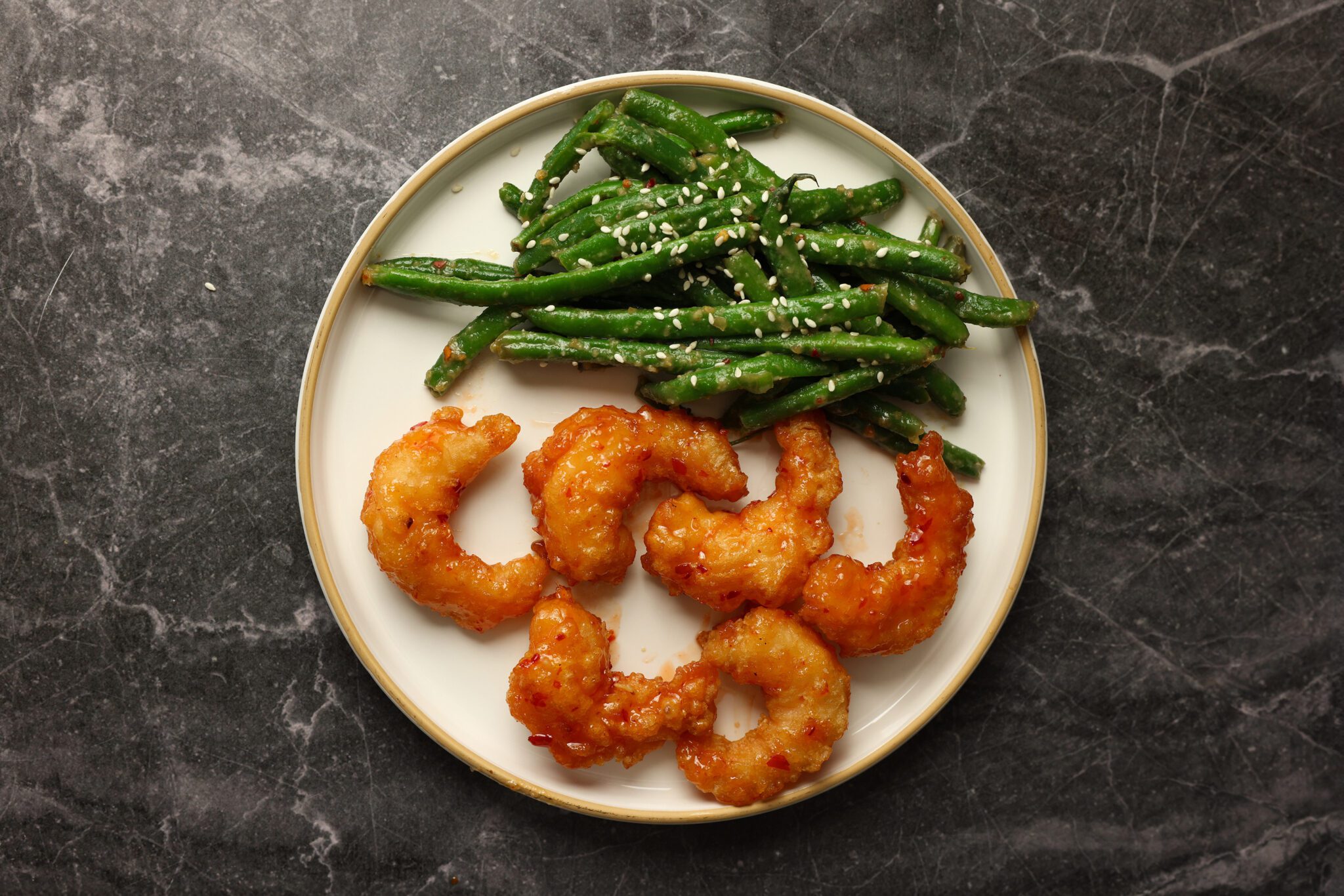 Overhead view of a white plate with sweet & spicy shrimp and miso green beans, served over a charcoal marble surface.