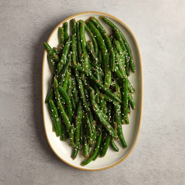 Overhead view of a white platter of miso green beans served over a white gray textured stone surface.