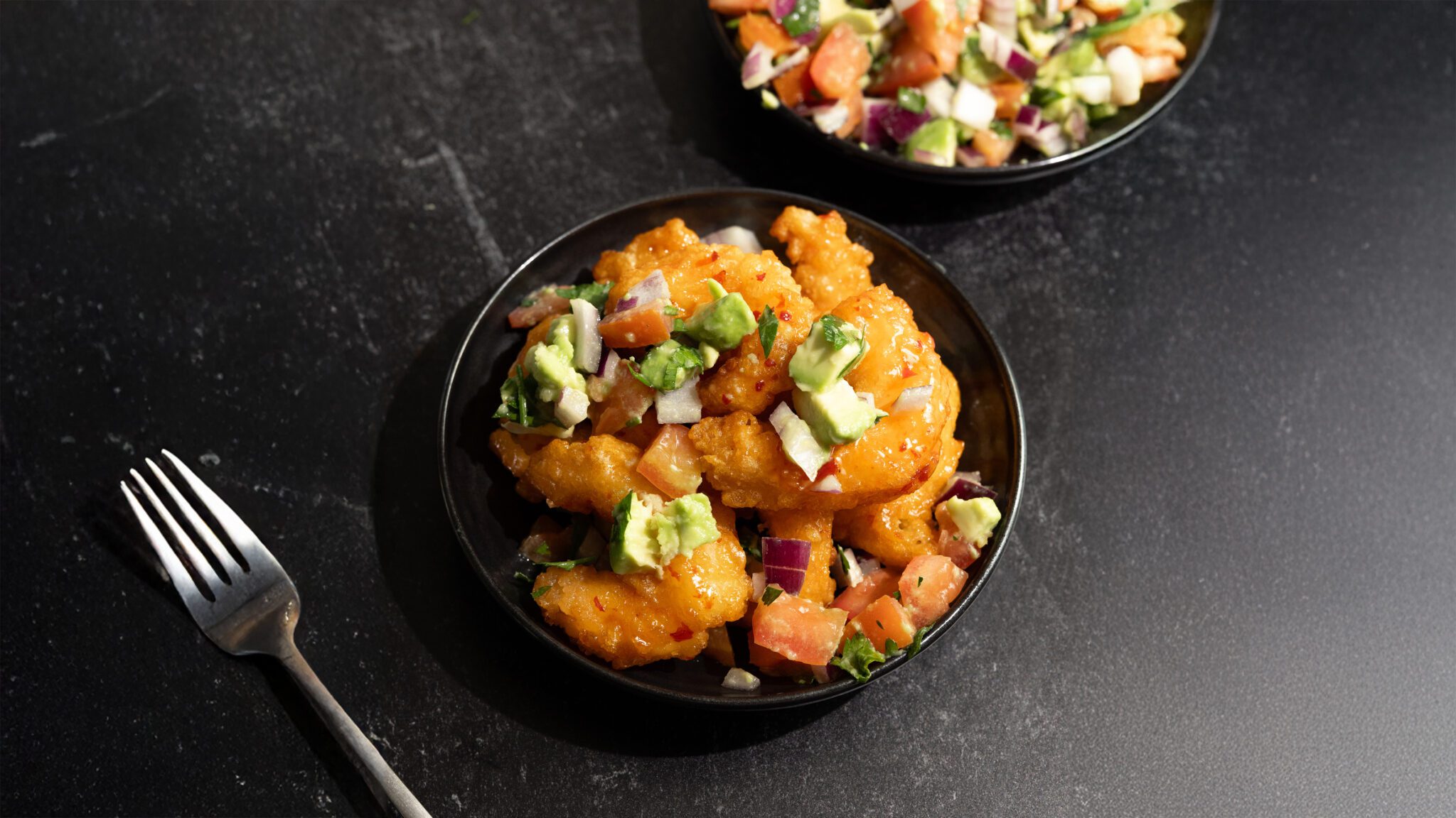 Three quarter view of Avocado Pico de Gallo and crispy shrimp in a black bowl and another bowl of pico de gallo. Both served over a black marble tabletop.