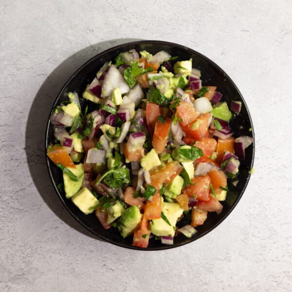 Overhead view of Avocado Pico de Gallo in a black bowl, served over a white textured tabletop.