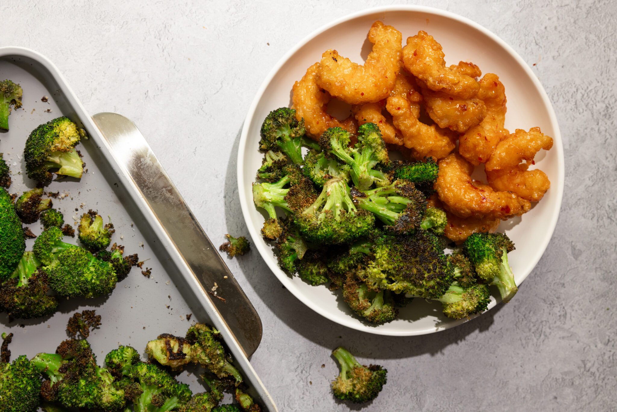 Overhead view of roasted broccoli on a sheet pan off to the side. On the right is a white plate of roasted broccoli and crispy sweet & spicy shrimp.