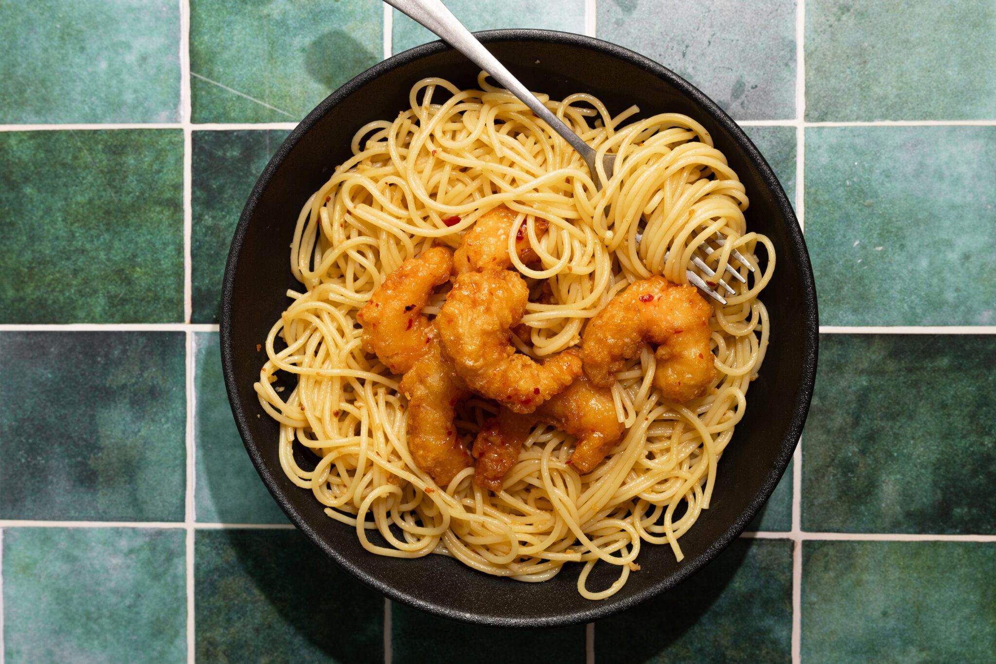 Overhead view of spicy garlic noodles with shrimp on a black plate with a fork, served over a green tiled surface.