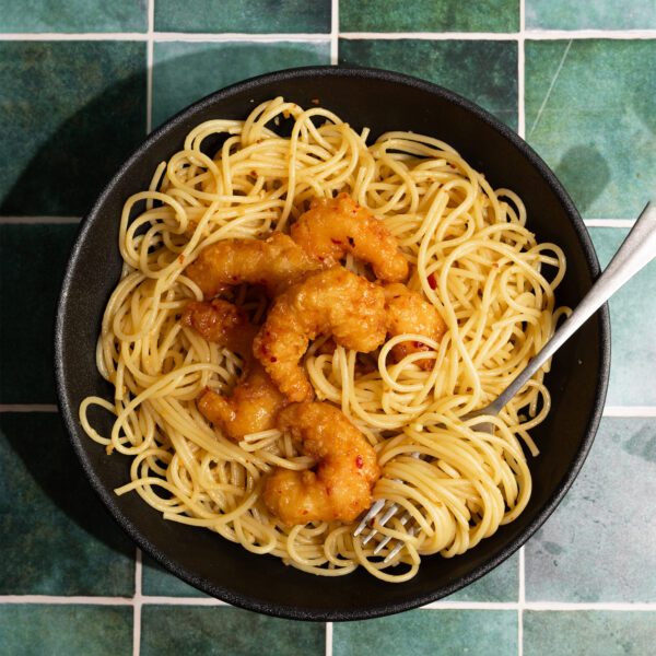 Overhead view of spicy garlic noodles with shrimp on a black plate with a fork, served over a green tiled surface.