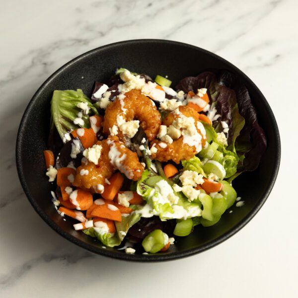 Three-quarter view of a black bowl of buffalo-inspired salad with crispy sweet & spicy shrimp, topped with blue cheese crumbles.