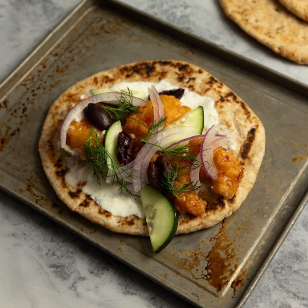 Three quarter view of a metal baking pan with a piece of pita bread, crispy shrimp, tzatziki, red onion, cucumber slices, sliced Kalamata olives, and fresh dill.