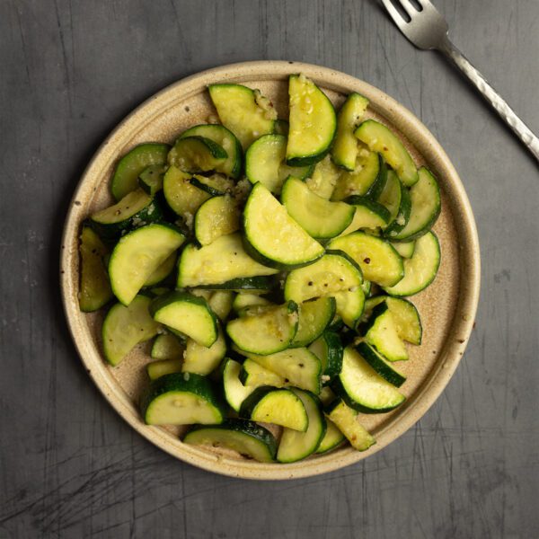 Overhead view of garlic zucchini on a beige plate over a textured charcoal tabletop with a silver fork off to the side.