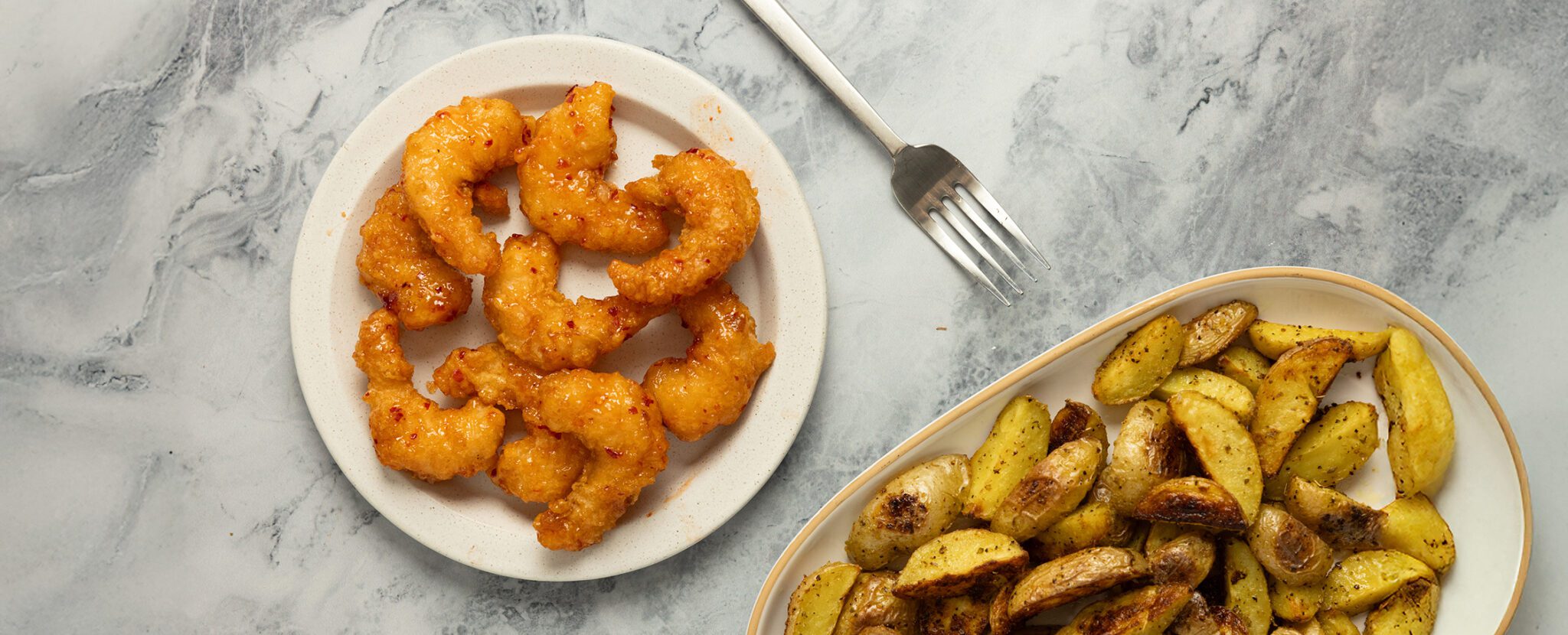 Overhead view of a round plate of sweet and spicy shrimp with a large platter of crispy adobo potato wedges and a sliver fork, all served over a marble tabletop.