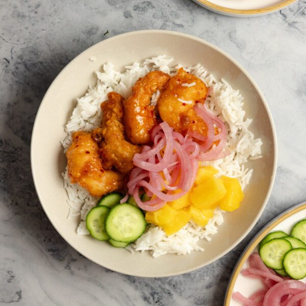 Overhead view of a white bowl of Social Kitchens Sweet & Spicy Shrimp, sliced cucumbers, pineapple chunks, pickled red onions, and white rice.