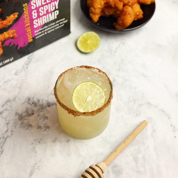 Three quarter angle view of Sweet & Spicy Margarita in a clear glass with a chili salted rim and lime slice garnish. On the marble table surface is also a wooden honey wand, a small black plate of sweet & spicy shrimp and a box of Social Kitchens Sweet & Spicy shrimp packaging.