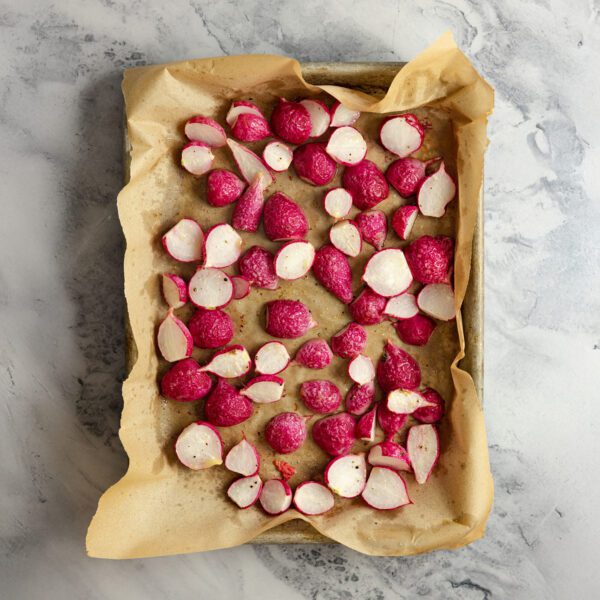 Overhead view of a sheet pan lined wtih brown parchment paper on a marble countertop. Filled inside the pan are sliced raddishes.