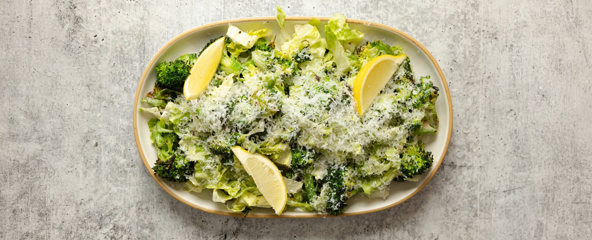 Overhead view of a large ceramic platter of Roasted Broccoli Caesar Salad with lemon wedges, served over a stone tabletop.