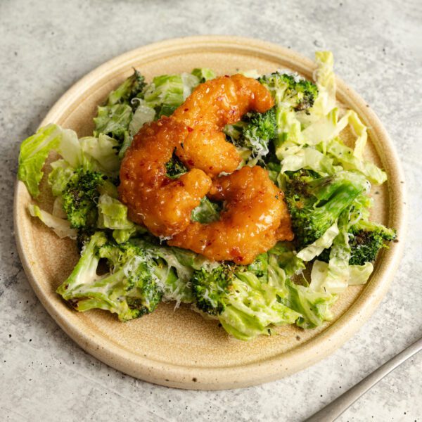 Three-quarter view of a beige ceramic plate of Social Kitchens Sweet & Spicy Shrimp over a bed of roasted broccoli caesar salad.
