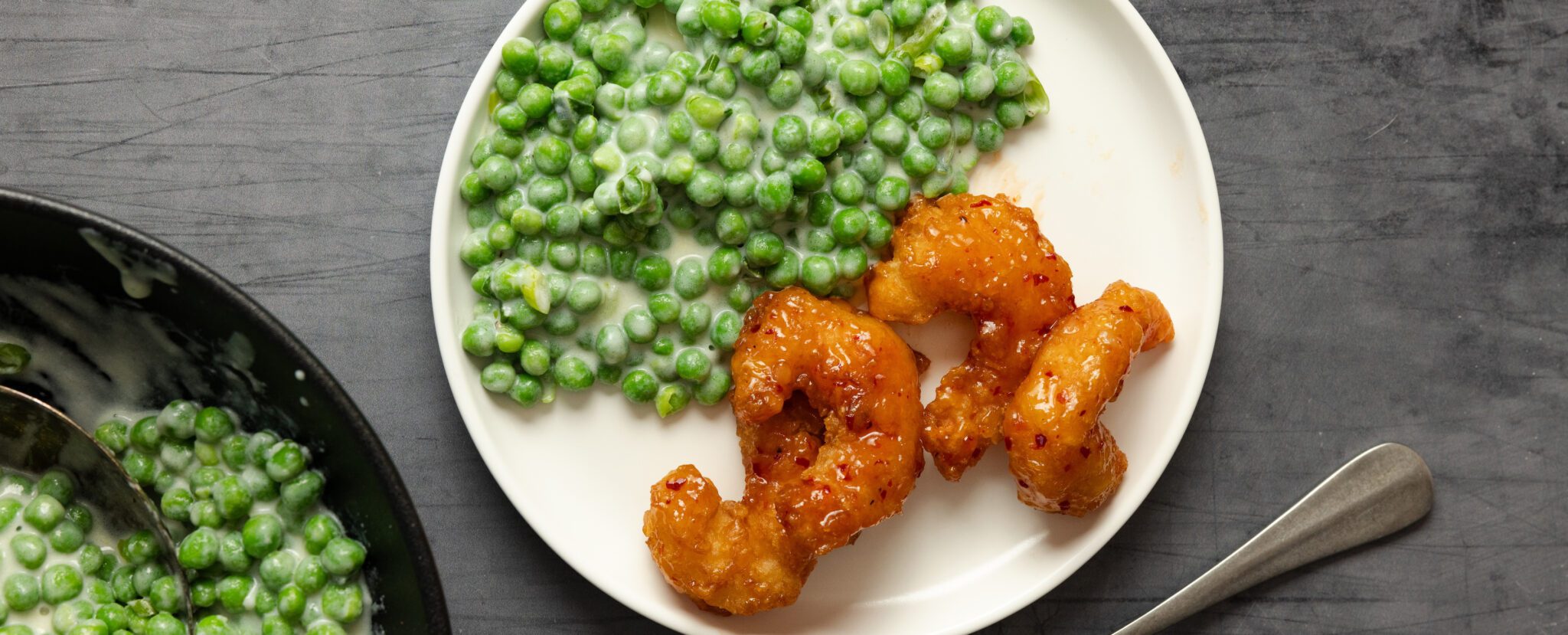 Overhead view of a white plate of Social Kitchens Sweet & Spicy Shrimp with a side of scallion creamed peas. Off to the side is another black bowl of the peas.