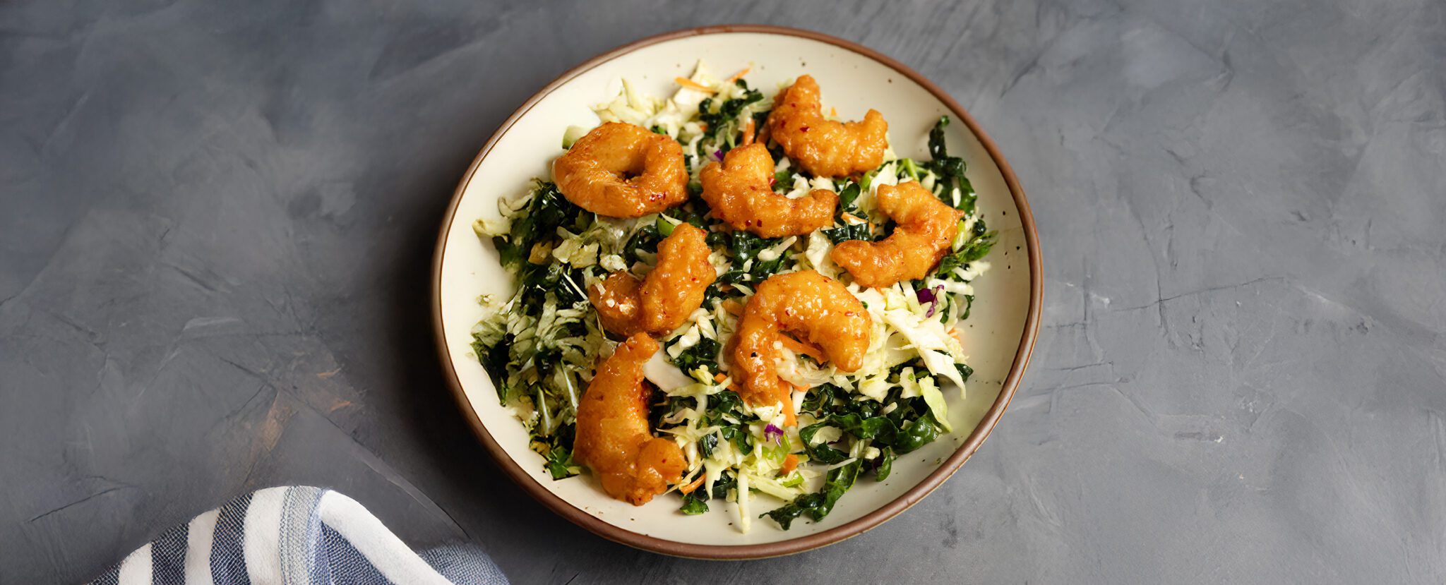 Three-quarter view of a white ceramic bowl of crunchy kale slaw with Social Kitchens Sweet & Spicy shrimp. Served on a gray textured tabletop with a white and gray striped napkin off to the side.