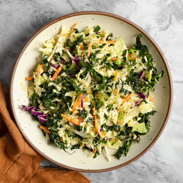Overhead view of white ceramic bowl filled with crunchy kale slaw. Served on a marble tabletop and a terracotta-colored linen napkin off to the side.