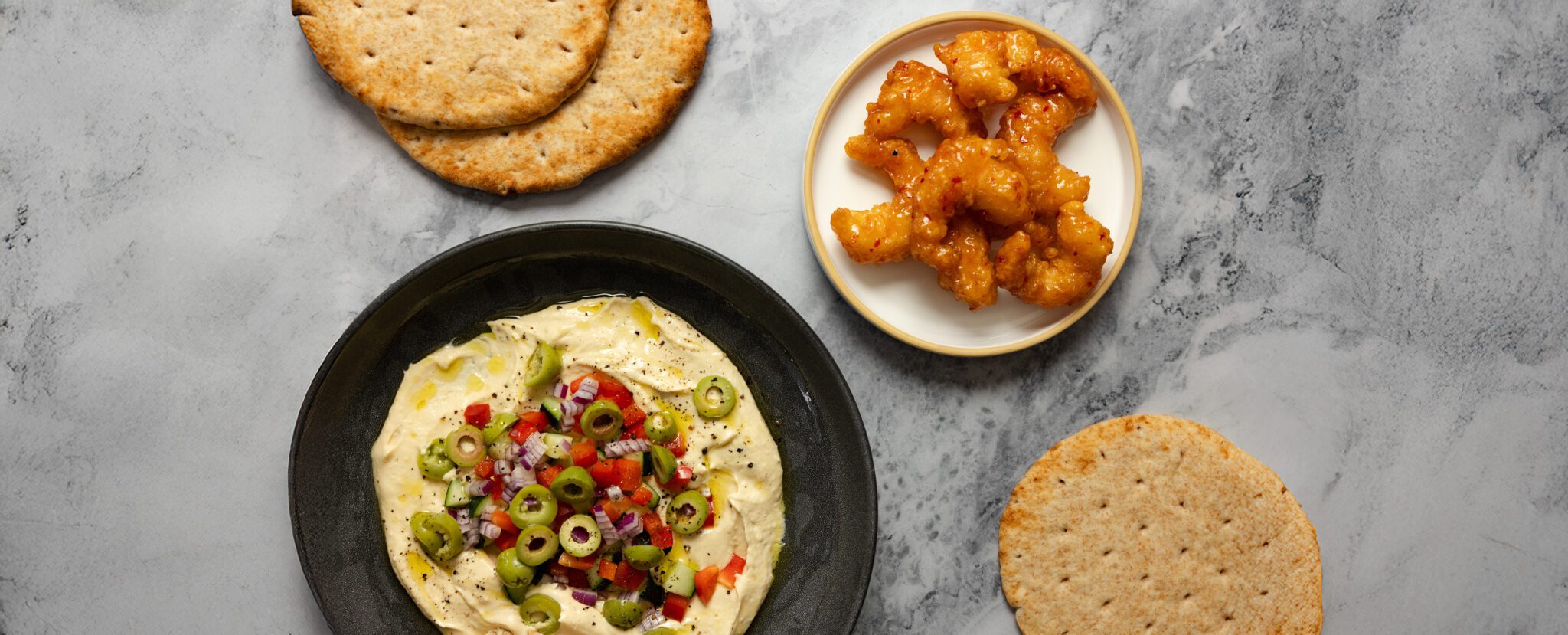 Overhead view of a white plate with shrimp, some pieces of pita bread, and a black bowl of hummus topped with olives, red bell pepper, red onion, and cucumbers.