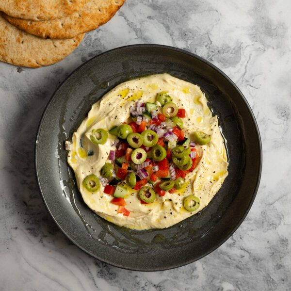Overhead view of a black bowl of hummus topped with olives, red bell pepper, red onion, and cucumbers with some pieces of pita bread off to the side.
