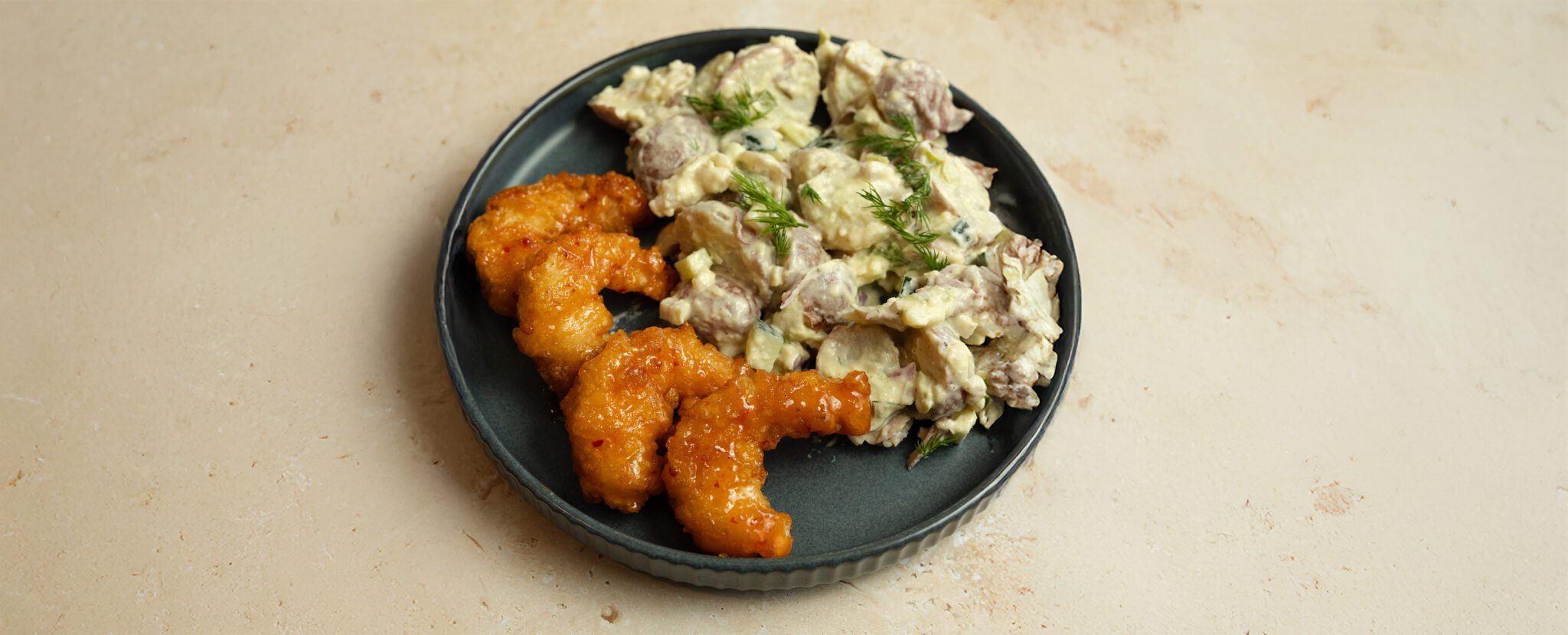 Three-quarter view of a black plate of Mediterranean Potato Salad and four pieces of Social Kitchens Sweet & Spicy Shrimp. Served over a beige marble tabletop.