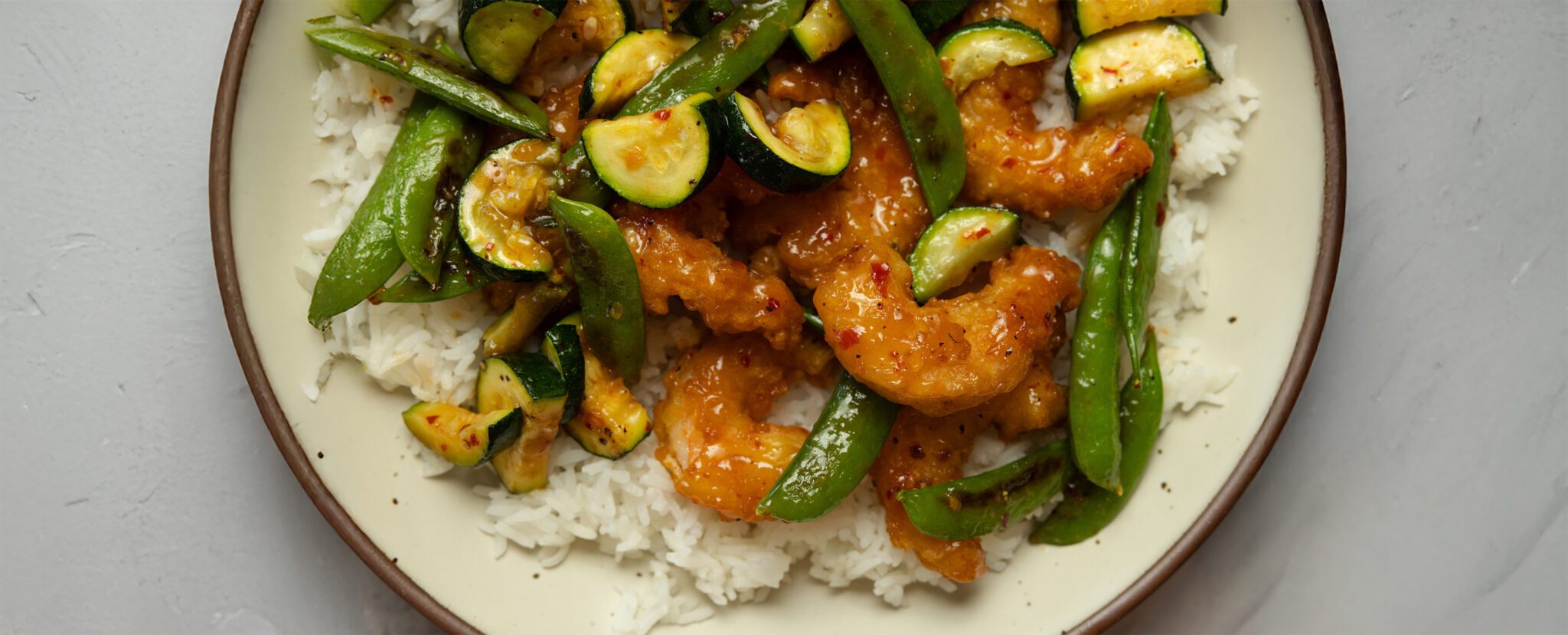 Cropped overhead view of summer veggies & shrimp sheet pan meal over rice on a white plate, served over a white marble tabletop.