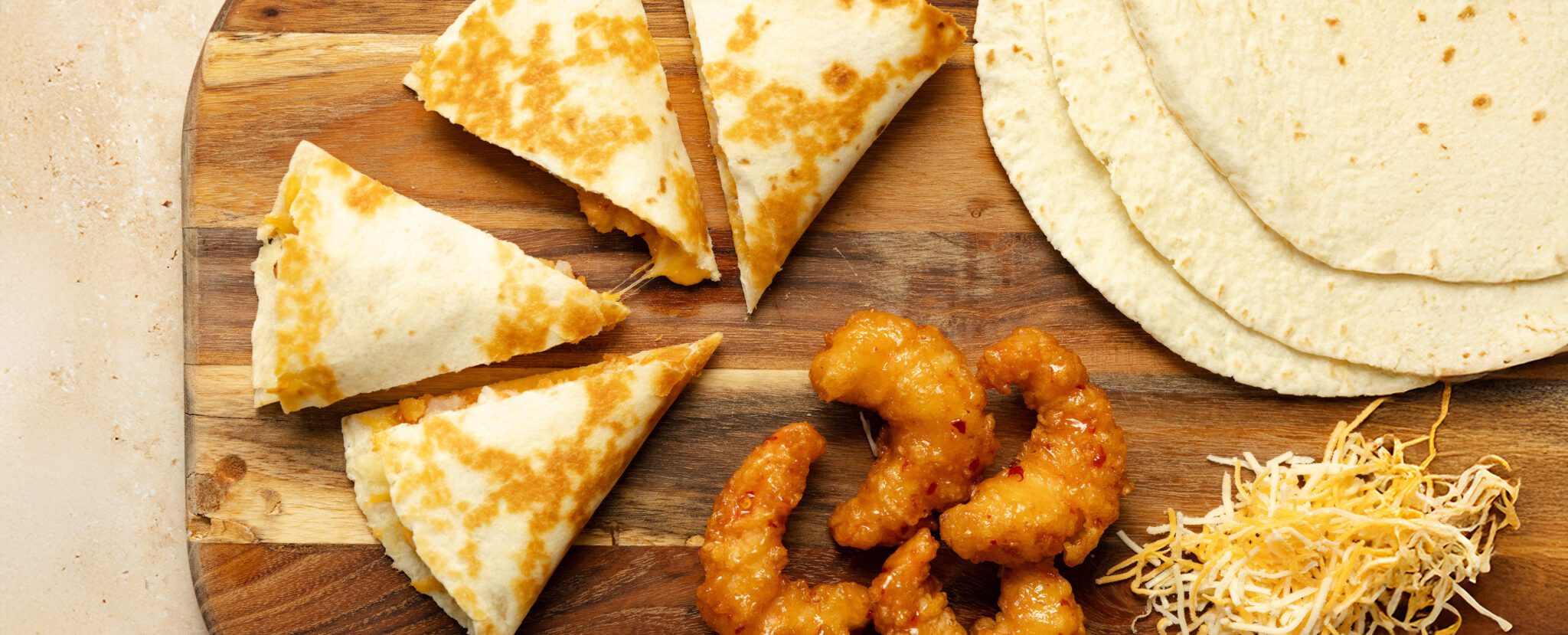 Overhead view of four quesadilla slices, 4 pieces of Social Kitchens Sweet & Spicy shrimp, several tortillas, and some shredded cheese all on a wooden cutting board.
