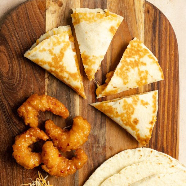 Overhead view of four quesadilla slices, 4 pieces of Social Kitchens Sweet & Spicy shrimp, several tortillas, and some shredded cheese all on a wooden cutting board.
